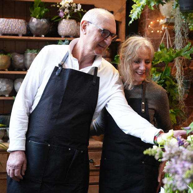 Two florists wearing handcrafted, black canvas aprons that are made in Britain with removable leather straps and pocket.