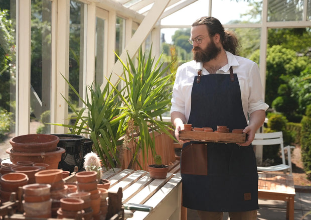 Male gardener wearing handcrafted, denim apron; made in Britain with removable, leather straps and pocket.